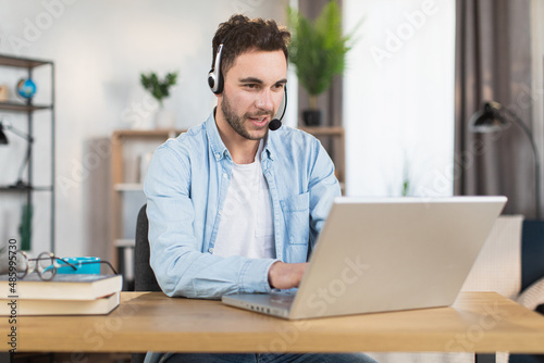 Young caucasian man in headset sitting at desk and having video call on modern laptop. Concept of gadgets, remote work and communication.