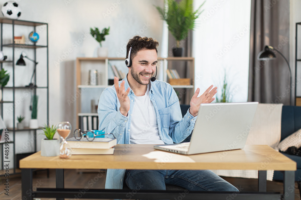 Handsome caucasian man talking and gesturing during video conference on modern laptop. Positive young guy using wireless headset while working at home.