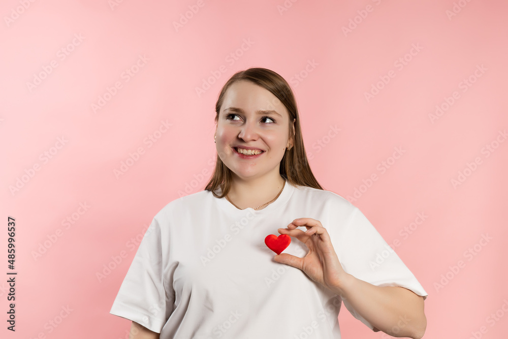Thoughtful young girl with brown hair in a white t-shirt on a pink background and red heart. High quality photo Thoughtful young girl with brown hair in a white t-shirt on a pink background and red heart. High quality photo