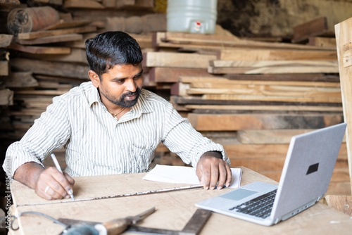 carpainter or woodworker measuring wood by seeing blueprint or plan on laptop at workplace - concept of technology, artisan and skilled labour. photo