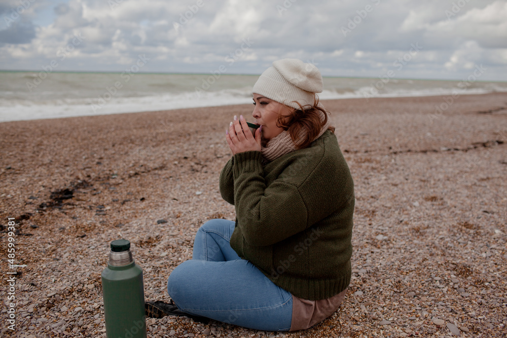 Beatuful woman with over weight body walking in the beach, and drink tea. The plus size model wearing jeans, the warm knitted blanket and sweater. the wind in the bich have fun with hairs