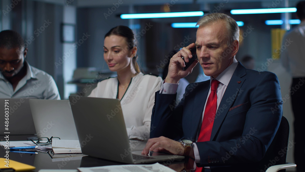 Mature businessman talking on cellphone and typing on laptop working with colleagues in office