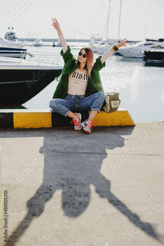 a young girl in jeans, a cardigan and sunglasses seets laughing by the sea against the background  yachts photo