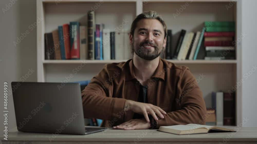 Caucasian male blogger waving hand and talking on camera while sitting at desk with portable laptop. Happy bearded man creating content for his vlog. Caucasian male blogger waving hand and talking on camera while sitting at desk with portable laptop. Happy bearded man creating content for his vlog.
