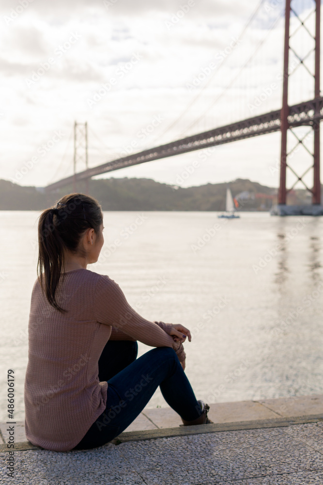 Unrecognizable young girl contemplating the 25 April Bridge sitting on the ground in Lisbon