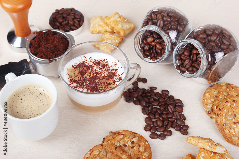 Breakfast with cup of espresso, cappuccino with chocolate crumbs, cookies, crackers, holder with ground coffee, tamper and coffee beans on table. Breakfast with cup of espresso, cappuccino with chocolate crumbs, cookies, crackers, holder with ground coffee, tamper and coffee beans on table.