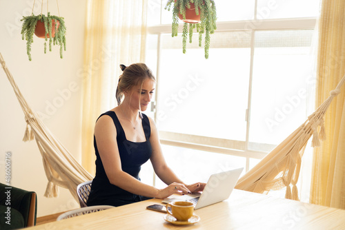 Young woman working at home in her kitchen with laptop and papers on kitchen wooden desk. Home office concept.