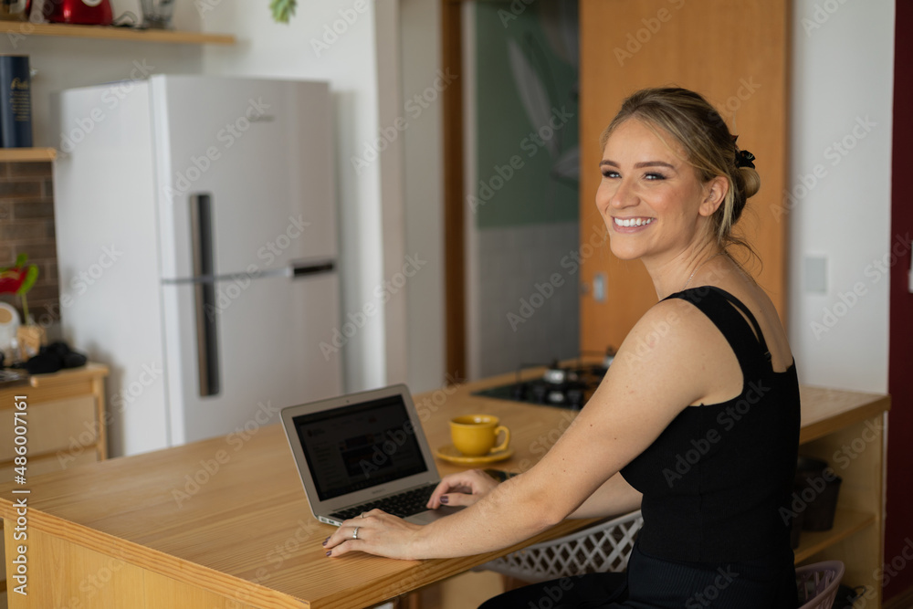 Young woman working at home in her kitchen with laptop and papers on kitchen wooden desk. Home office concept. Young woman working at home in her kitchen with laptop and papers on kitchen wooden desk. Home office concept.