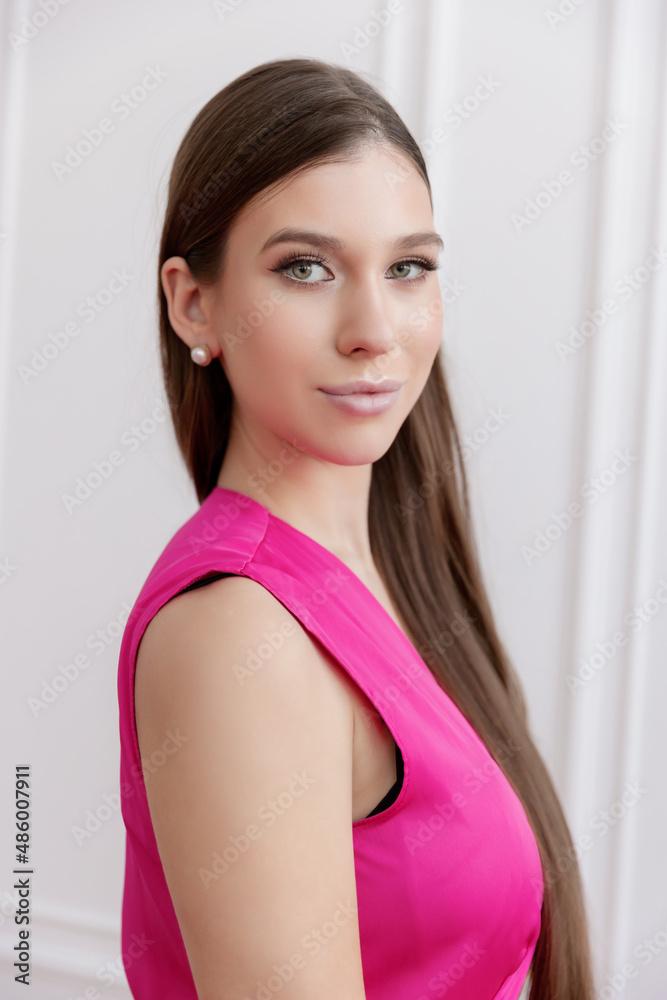 Portrait of a young brunette Caucasian woman with long straight hair in a bright pink dress. Revealed thigh-high slit. In the background is a white wall and a fireplace Portrait of a young brunette Caucasian woman with long straight hair in a bright pink dress. Revealed thigh-high slit. In the background is a white wall and a fireplace