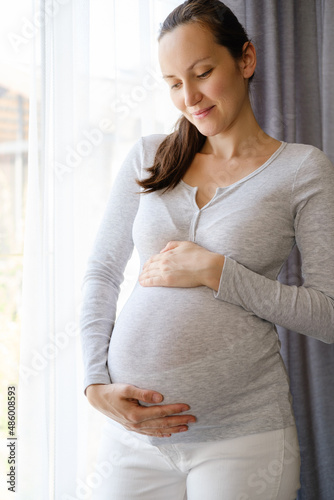 caucasian happy pregnant woman with hands on belly standing near window
