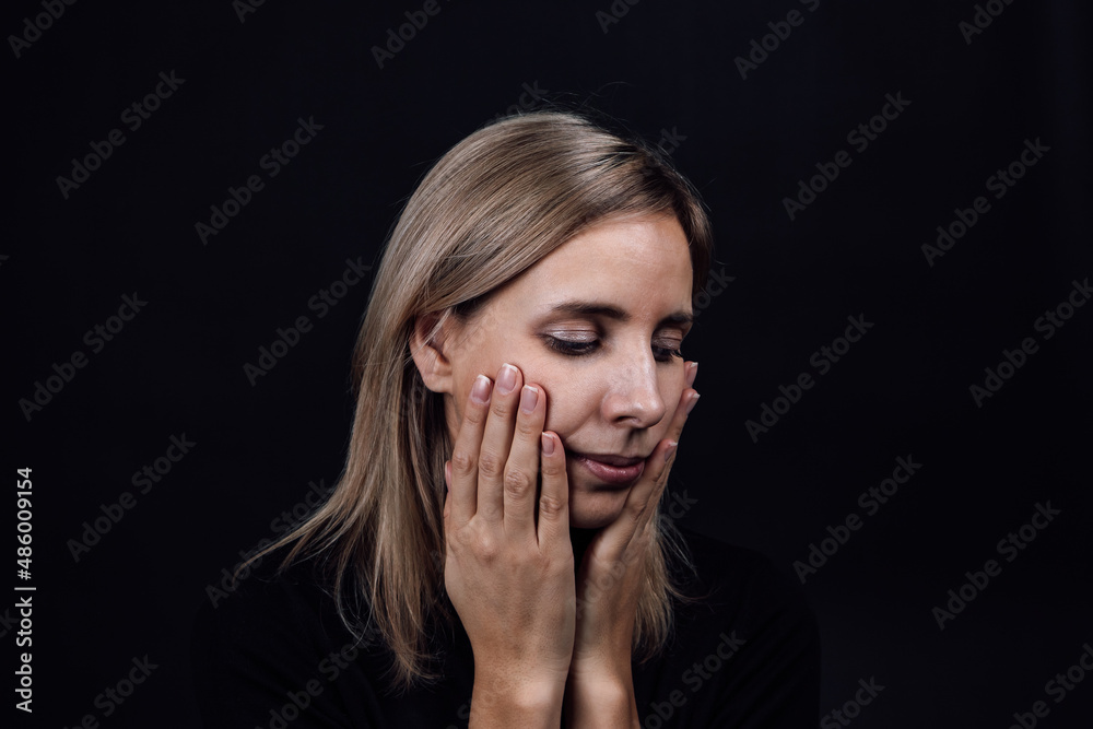 Depressed woman face with hands touching cheeks wearing black dress on black background. Victim of physical and psychological abuse. Gaslighting. Relative aggression.