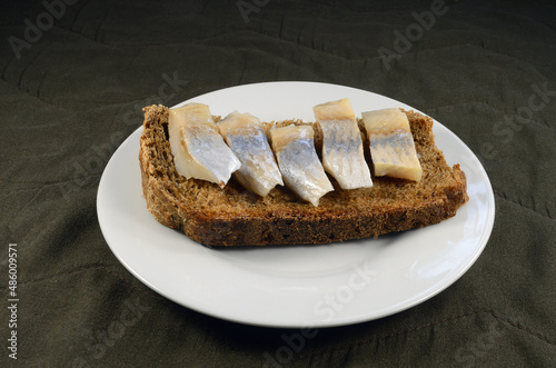 Fresh rye bread sandwich with herring on a white saucer, brown fabric background.