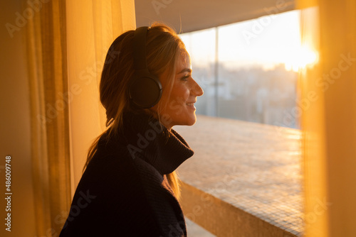 Side view of young woman listening to music with headphones in her ears by the window with a city view landscape in the background. High quality photo © Buonaventura