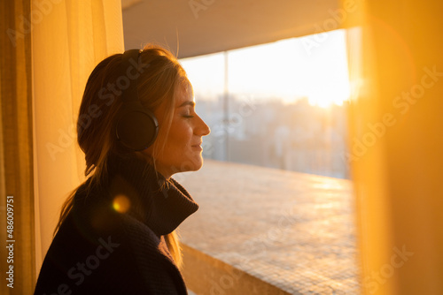 Side view of young woman listening to music with headphones in her ears by the window with a city view landscape in the background. High quality photo