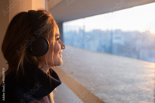 Side view of young woman listening to music with headphones in her ears by the window with a city view landscape in the background. High quality photo