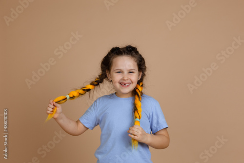 Funny, cheerful joyful female child with missing tooth smiling looking at camera holding yellow kanekalon braids with hands on beige background wearing blue t-shirt and jeans.