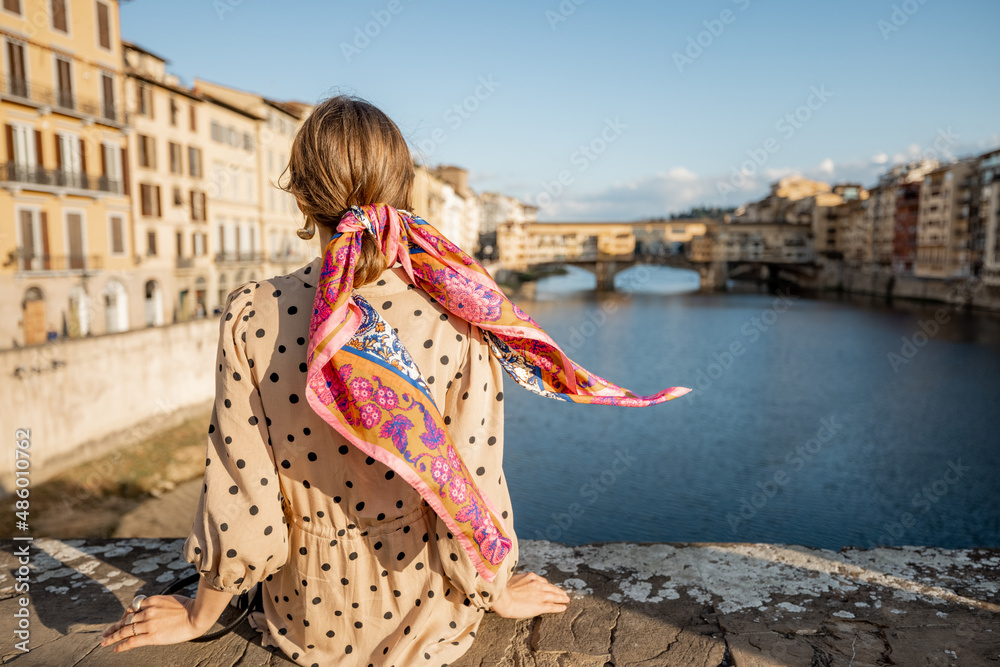 Fototapeta premium Young woman enjoys beautiful view on famous Old bridge in Florence, sitting back on the riverside at sunset. Female traveler visiting italian landmarks. Stylish woman wearing dress and colorful shawl