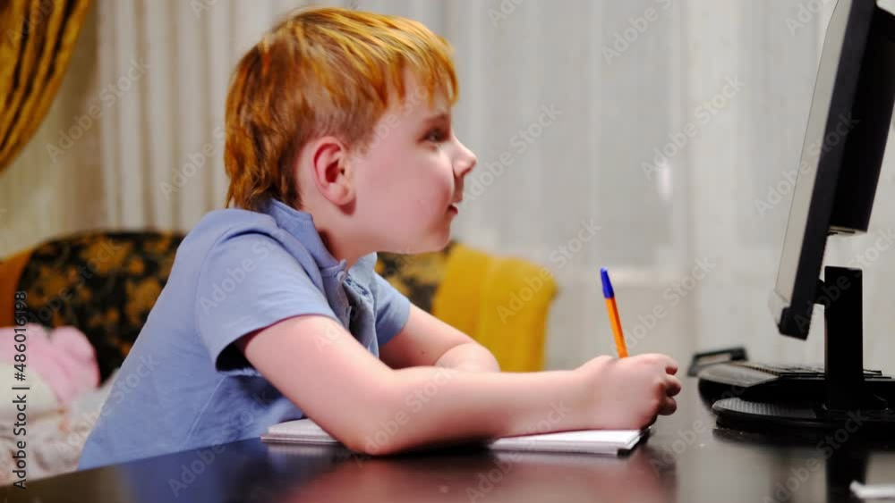 A young boy does homework, studies remotely, writes in a notebook in front of a computer screen. Home schooling. Quarantine. A young boy does homework, studies remotely, writes in a notebook in front of a computer screen. Home schooling. Quarantine.