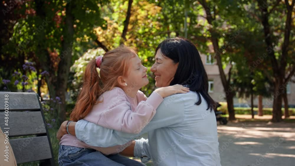 Senior woman squatting and hugging granddaughter outdoors on bench in park.