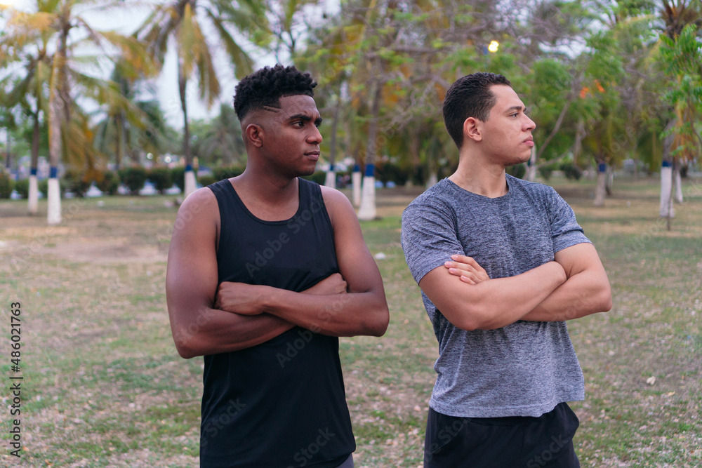 Two men in sportswear, standing in a park, smiling with their arms crossed. Two men in sportswear, standing in a park, smiling with their arms crossed.