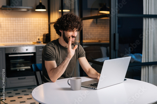 Cheerful young bearded man waving hand hello and talking to friends via video call using laptop sitting at table in kitchen room with modern interior. Happy male talking on video conference online.