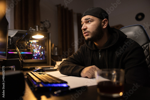 Portrait of a man addicted to computers, working remotely in IT industry, infromatics, programmer, hacker, internet criminal, mysterious guy in black sweatshirt with baseball cap, dark room