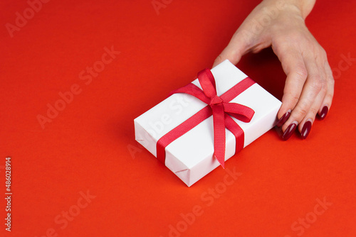 Woman hand hold white gift box with red bow on red table. Valentines day or birthday prsent concept