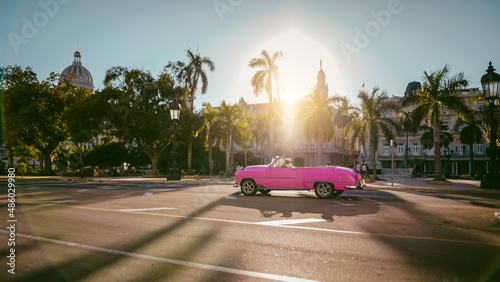 Havana, Cuba. Różowy cabriolet w świetle zachodzącego słońca.  Jedzie ulicą miasta pod palmami i niebieskim niebem photo