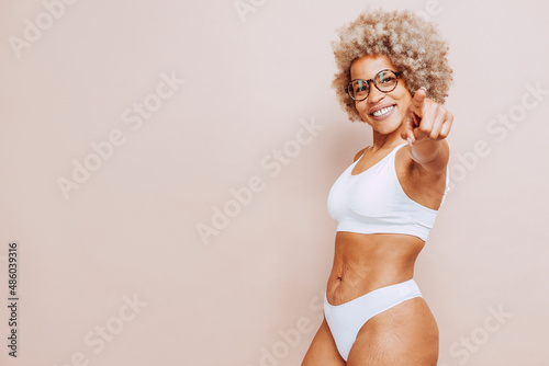 Cheerful beautiful afro latin american woman in white underwear and eyeglasses pointing a finger at camera, isolated on beige background at studio. Real body, natural beauty.