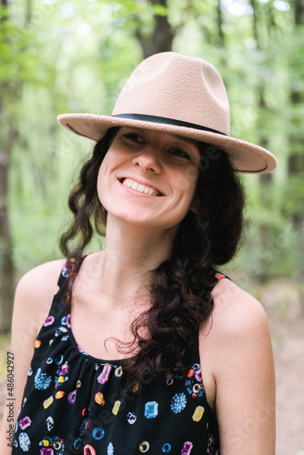 Young millennial woman in dress and beige hat stands in forest happy and relaxed, enjoys nature and tranquility. Travel alone concept.
