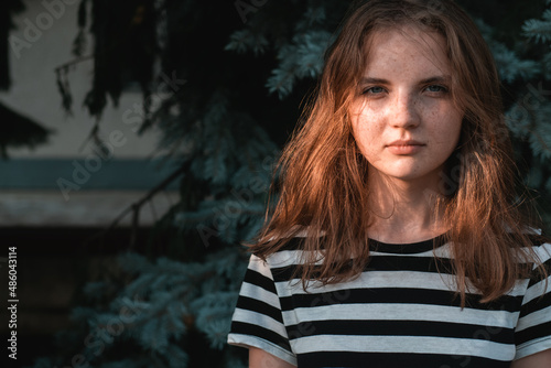 Portrait of young lovely woman with blue eyes and freckles in striped T-shirt and jeans against background of green foliage.