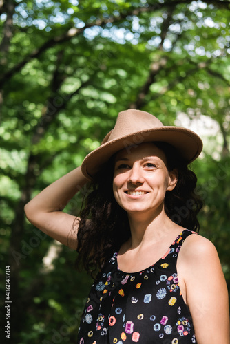 Young millennial woman in dress and beige hat stands in forest happy and relaxed, enjoys nature and tranquility. Travel alone concept.