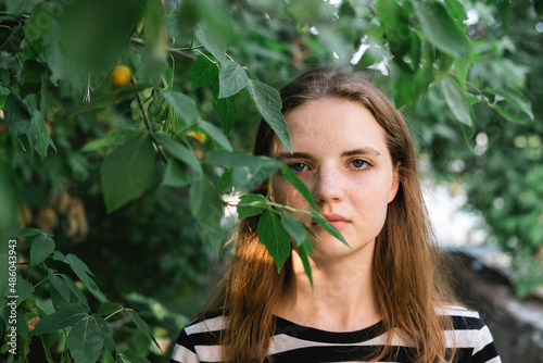 Portrait of young lovely woman with blue eyes and freckles in striped T-shirt and jeans against background of green foliage.