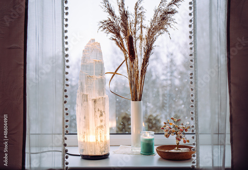 Rough big selenite crystal tower pole lamp illuminated on home window sill, spiritual home decor accent. Winter forest on background. photo