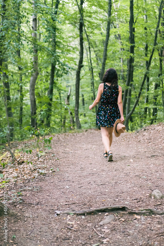Young millennial woman in dress and beige hat stands in forest happy and relaxed, enjoys nature and tranquility. Travel alone concept.