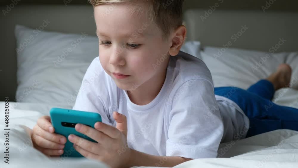 Cute little boy toddler playing mobile game, lying on a bed and holding smartphone Cute little boy toddler playing mobile game, lying on a bed and holding smartphone