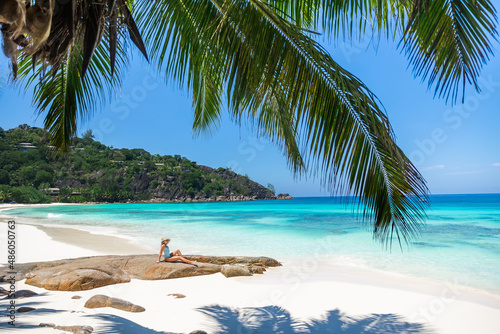 Young attractive woman posing on a stone in a blue swimsuit enjoying relaxing on a tropical beach with water, squeak and palm trees in the foreground