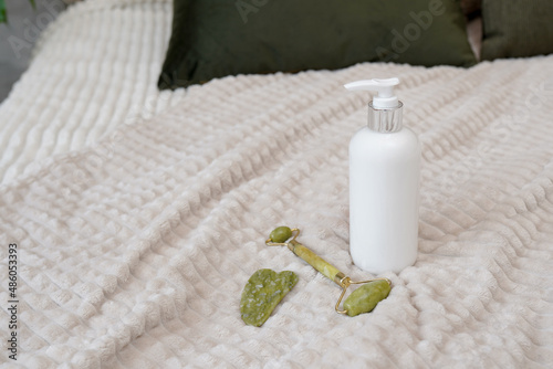 Set for home face care on the bed. Facial green quartz massage roller or gua sha and сosmetic white bottle of moisturizing lotion for skin care. Cosmetic