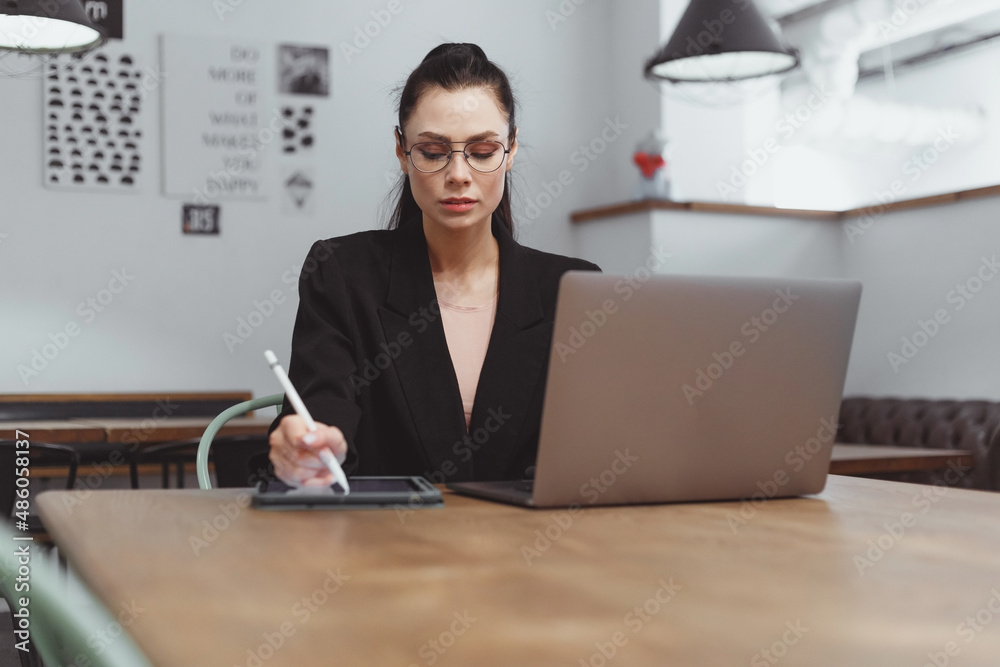 Young brunette woman in black suit drawing on tablet and work on laptop in empty cafe Young brunette woman in black suit drawing on tablet and work on laptop in empty cafe