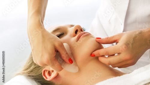 Beautician cosmetologist makes lifting modeling face massage with Gua Sha quartz scraper, professional cosmetic procedure in a beauty clinic salon for client. Skin care and cosmetology spa concept.