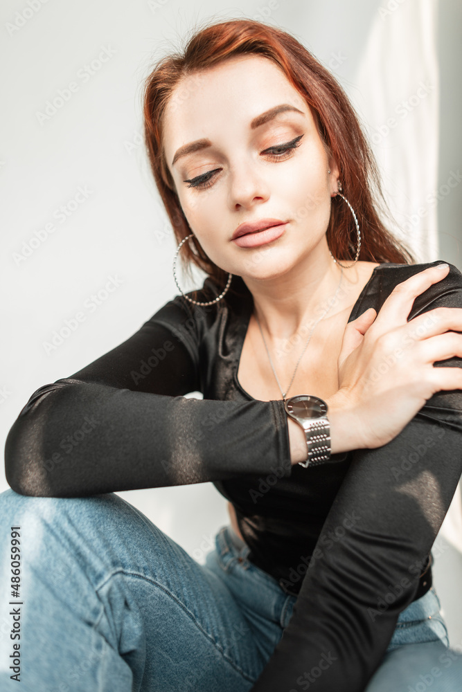 Fashion young woman model with cute face and clean skin in stylish black t-shirt and blue jeans sits indoors at sunlight Fashion young woman model with cute face and clean skin in stylish black t-shirt and blue jeans sits indoors at sunlight