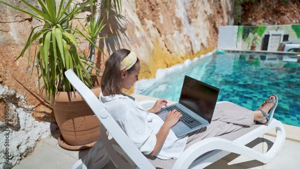 camera moving behind and around Happy woman enjoying relaxing by the pool with laptop computer with white empty screen browsing online during vacation at resort hotel.
