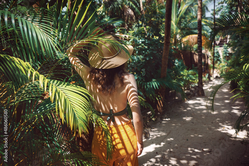 Young beautiful woman stands in the shade of palm trees, view from the back. A girl walks among tropical greenery on a bright sunny day in Mexico, Tulum photo