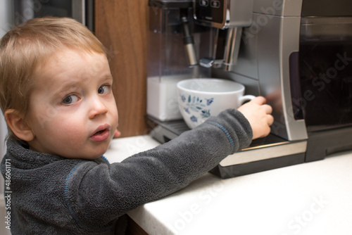 Little kid in kitchen. Danger for children at home. Baby and caffee machine. Little child try to grab cup of hot coffee. Attention of burn.