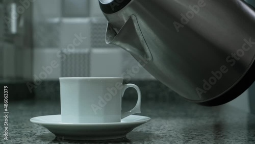Pouring boiling water from an electric kettle into a white mug standing on the kitchen table