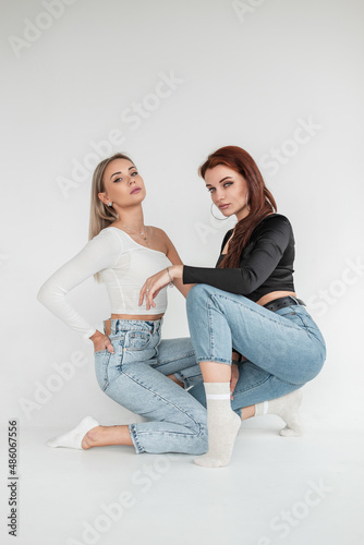 Two beautiful young fashionable girls in stylish denim clothes with T-shirts and bare vintage jeans in the studio sits and poses on a white background