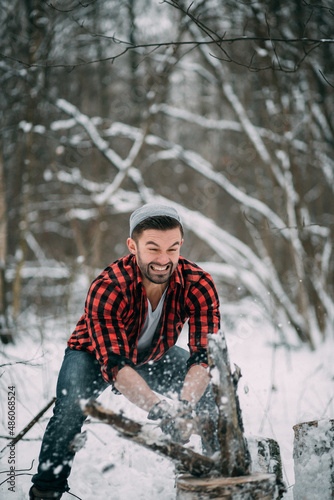 A male lumberjack is chopping firewood with an axe in a winter forest. A bearded man in a plaid shirt, beige vest and jeans. Chopping wood with all my might, face tense