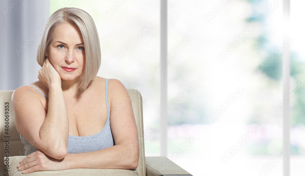 Beautiful middle-aged blonde woman sitting relaxed near the window. Selective focus on face. Realistic images with their own imperfections.