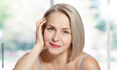 Happy beautiful middle-aged blonde woman shows off her perfectly well-groomed face and sitting relaxed near the window. Realistic images.