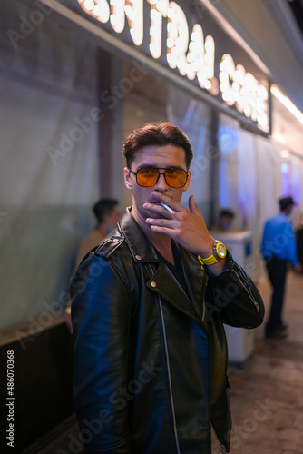 Portrait of young handsome brunet caucasian man in black leather jacket and yellow sunglasses. Standing on the street at night and smoking.